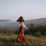A woman in a flowing skirt walks through a misty meadow with hills in the background.